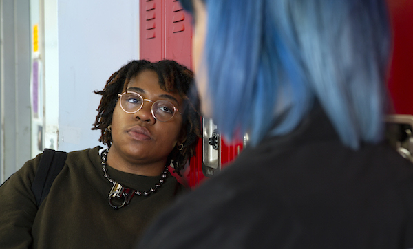 A non-binary student talking to a friend in front of their locker.