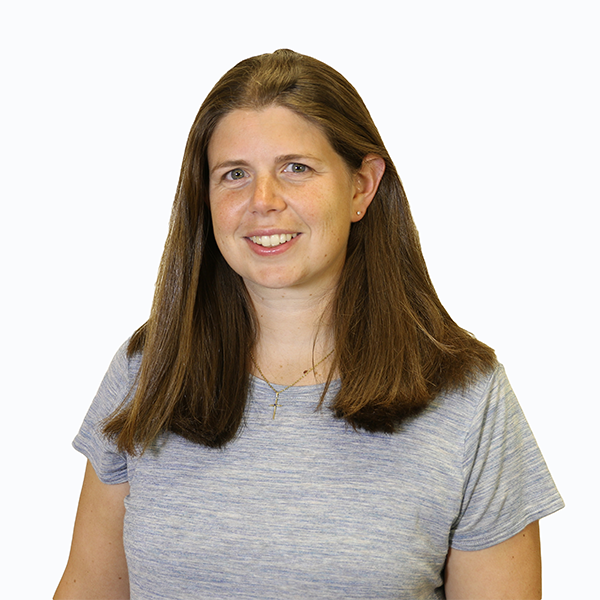Emily Sisson, with shoulder-length, straight brown hair, wearing a grey top, stands smiling at the camera