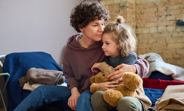 Young woman giving hug to her toddler son with brown soft teddybear while both sitting on sleeping place prepared for refugees