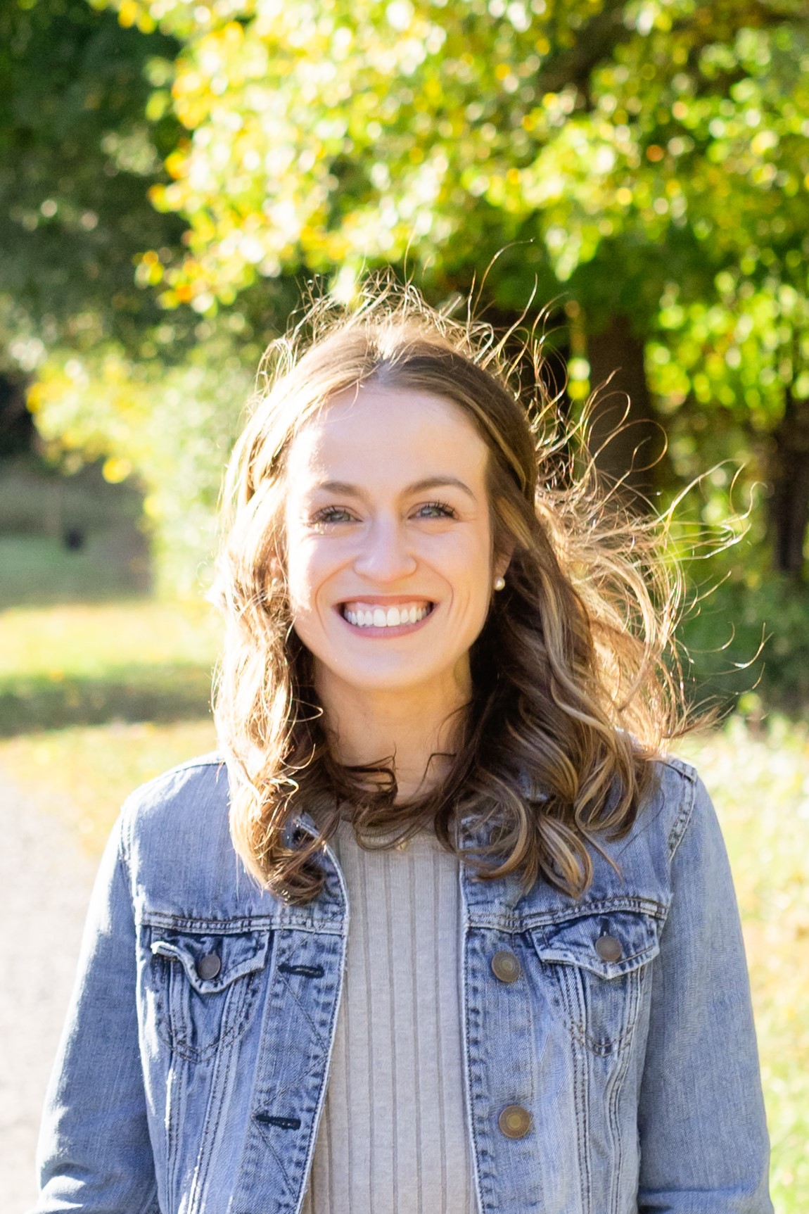 Headshot of Brittany Loewenstein smiling, wearing a jean jacket and grey shirt, and standing in a field in front of some trees