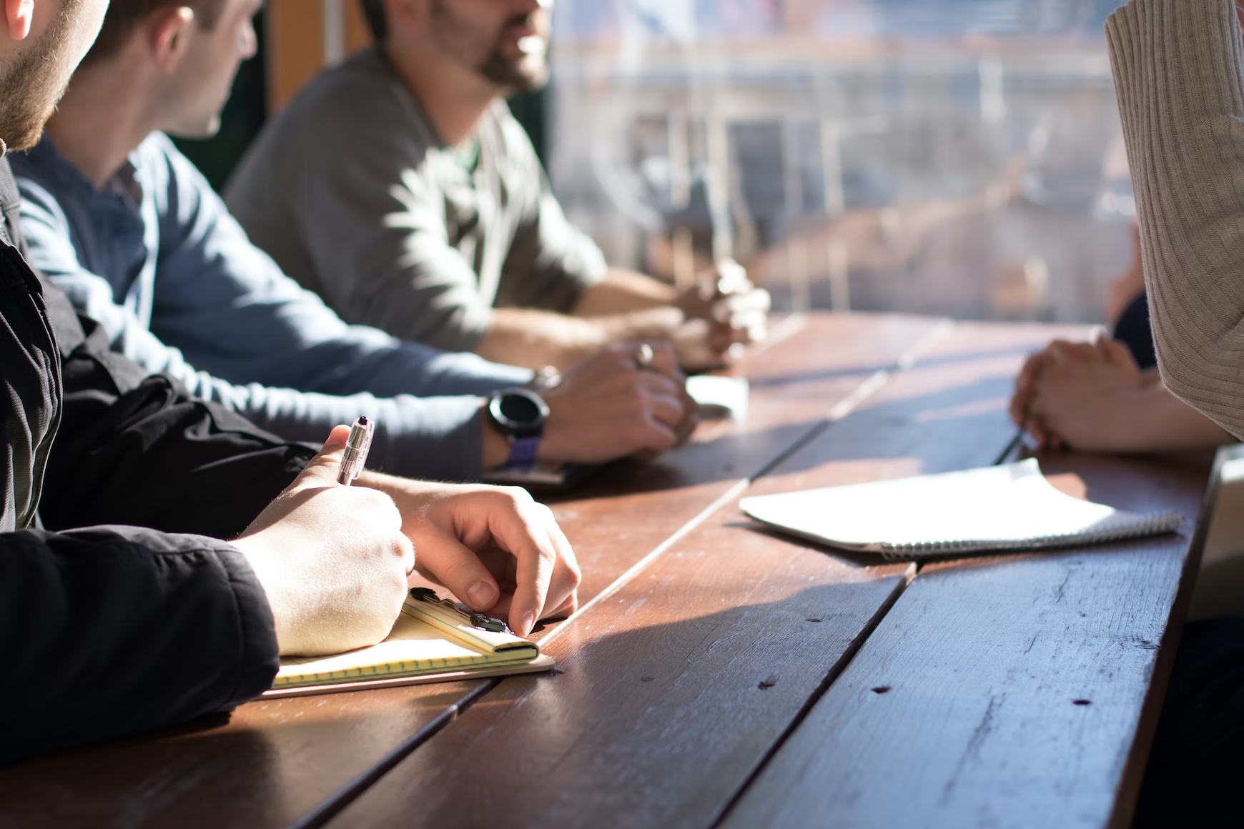 Four people sit at a wooden table with notepads