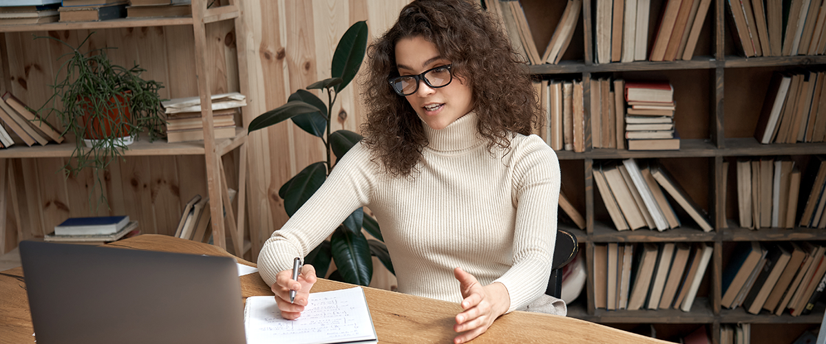 Woman in glasses speaking to a laptop, takes notes in front of a bookcase
