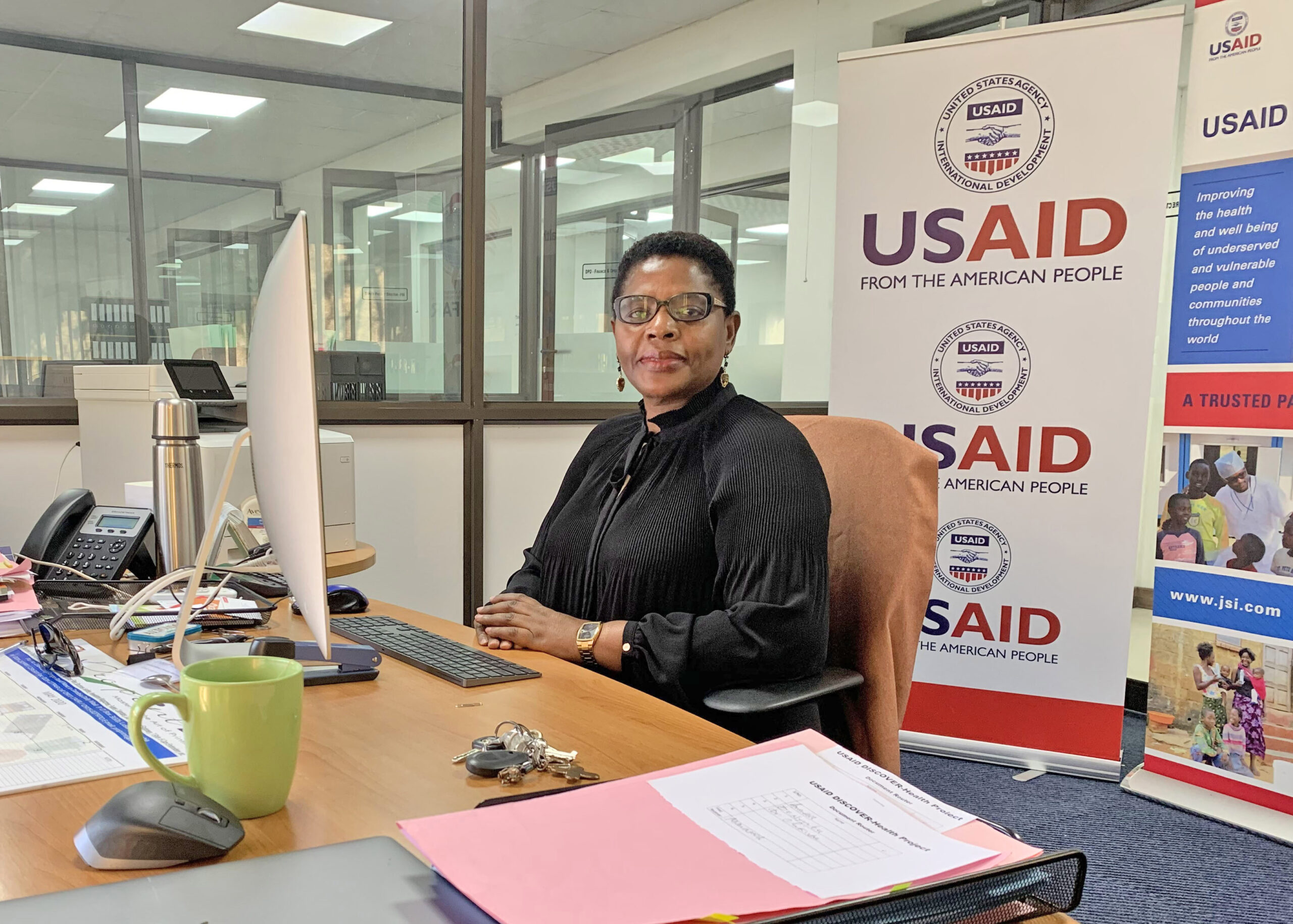 Muka Chikuba-McLeod sits at a desk in front of a USAID sign