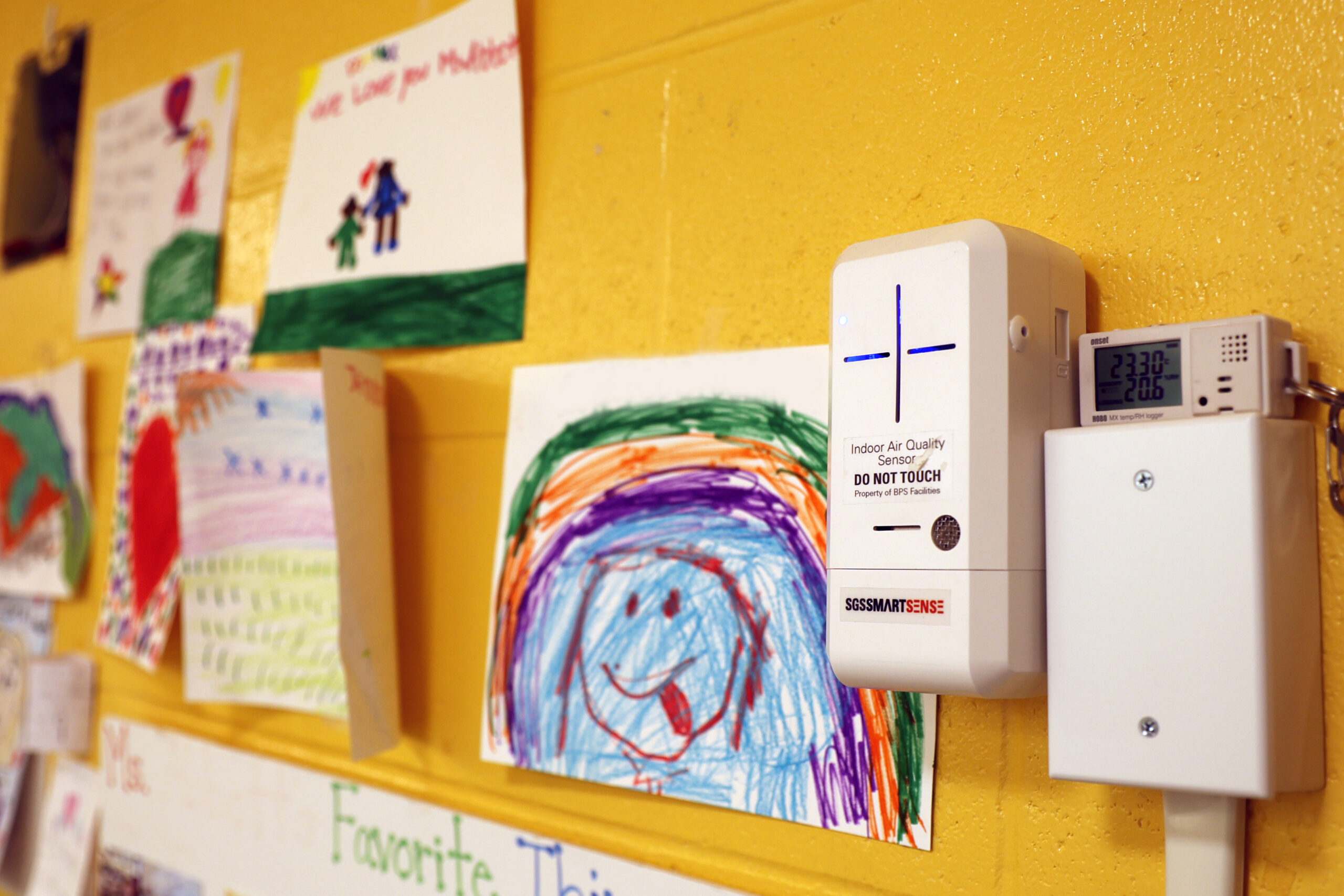An indoor air quality sensor installed on a yellow wall surround by young children's artwork