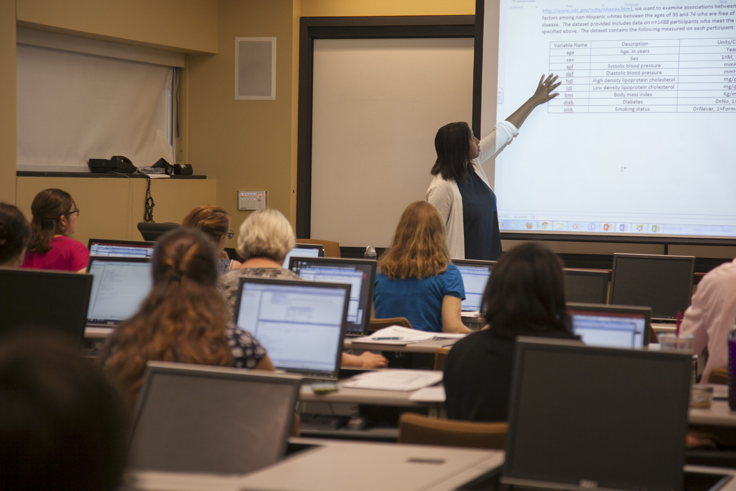 Jacqueline Hicks points to a slide projected at the front of a classroom, students sit with their laptops at rows of desks