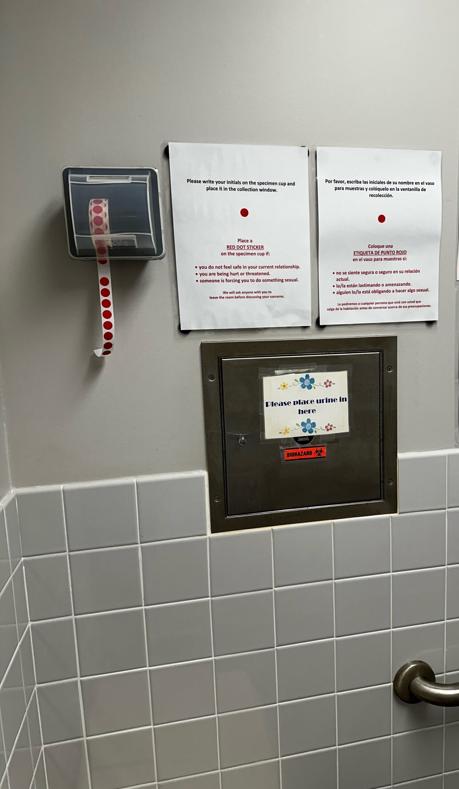 A roll of red circle stickers hangs from a box on a bathroom wall. One sticker is missing.