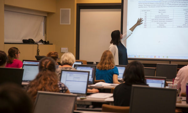 Jacqueline Hicks points to a slide projected at the front of a classroom, students sit with their laptops at rows of desks