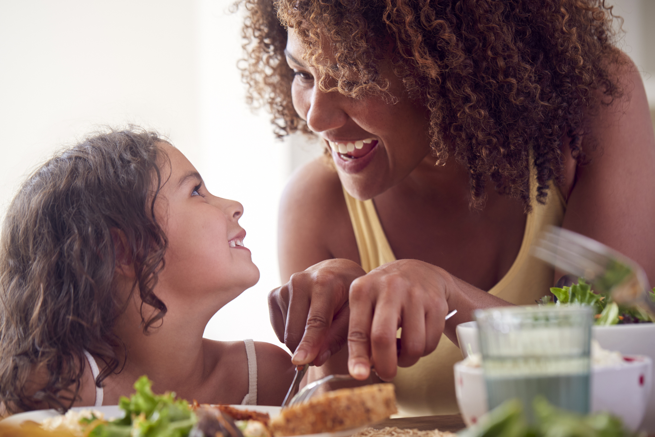 Mother Helping Daughter To Cut Food Sitting Around Table At Home Eating Family Meal Together