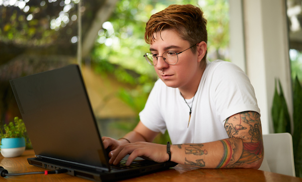 Transgender freelancer works on laptop in tropical coworking space. Focused individual with tattoos tech for remote job. Open, inclusive work plants. Creativity, productivity LGBTQ-friendly setting.