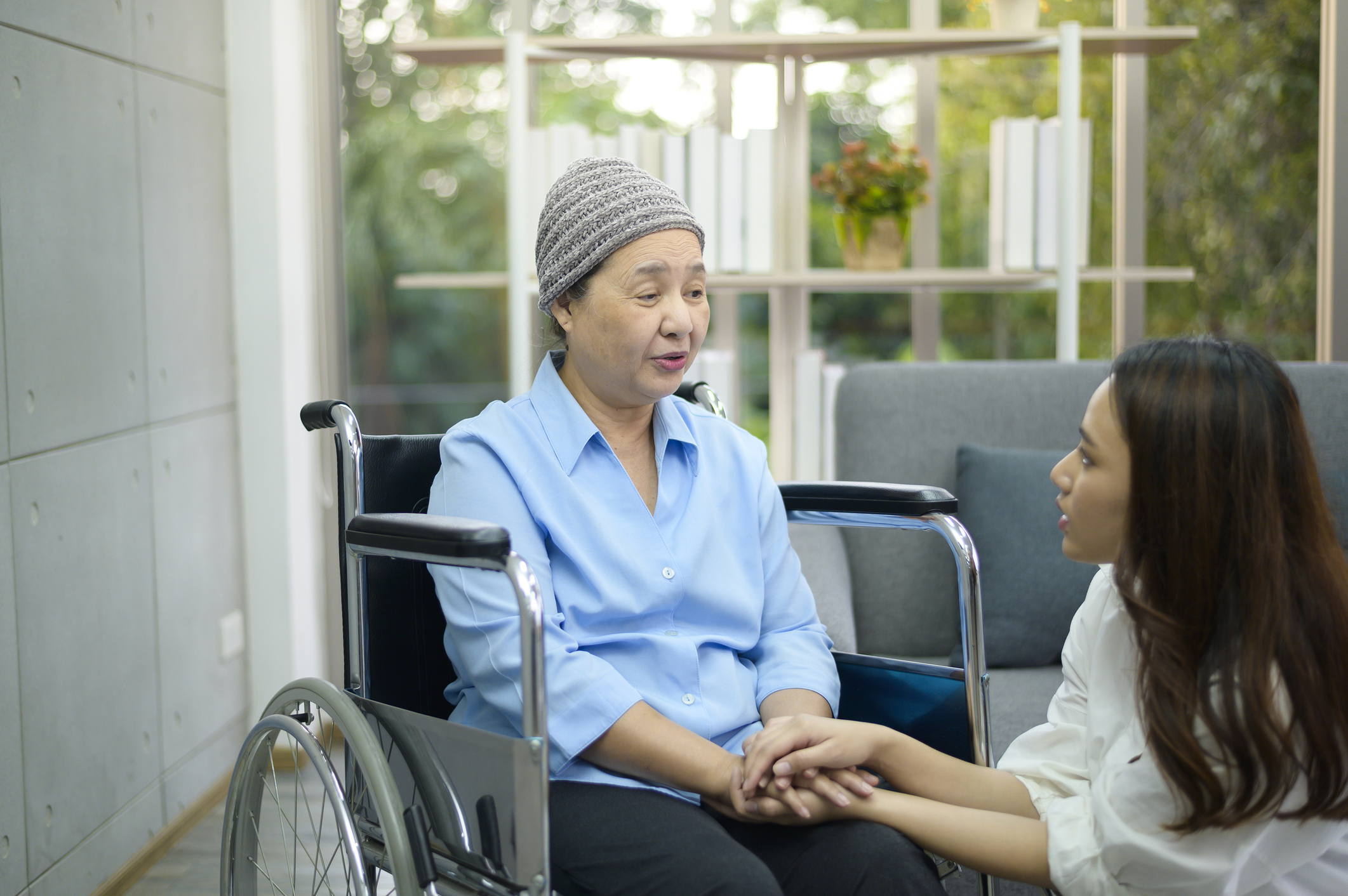 Cancer patient woman wearing head scarf sitting on wheelchair talking to her supportive daughter indoors, health and insurance concept.