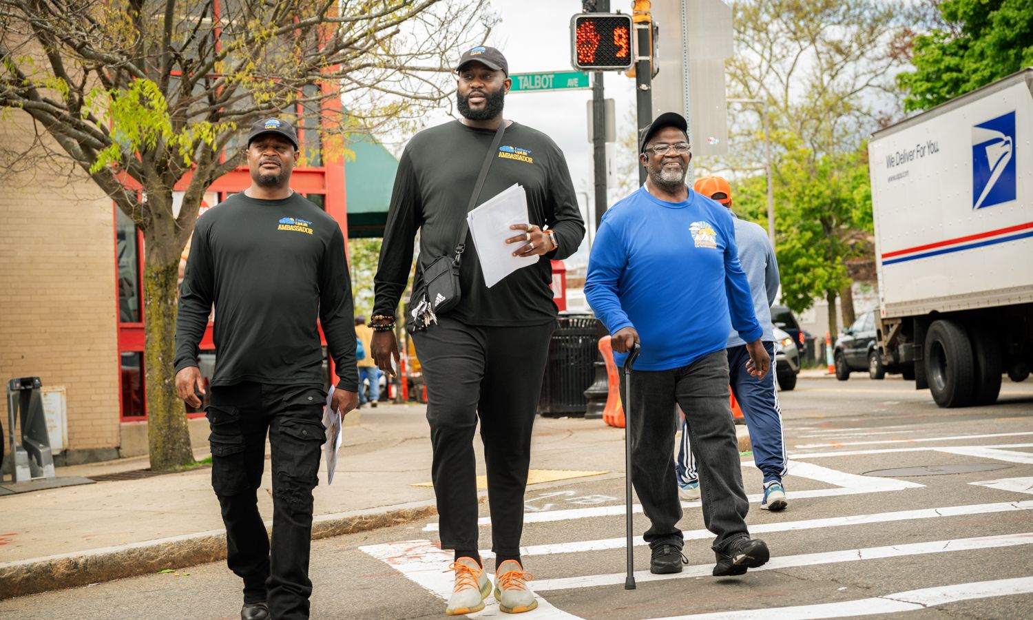 3 Black men cross the street wearing t-shirts with "Fathers Uplift" on them.