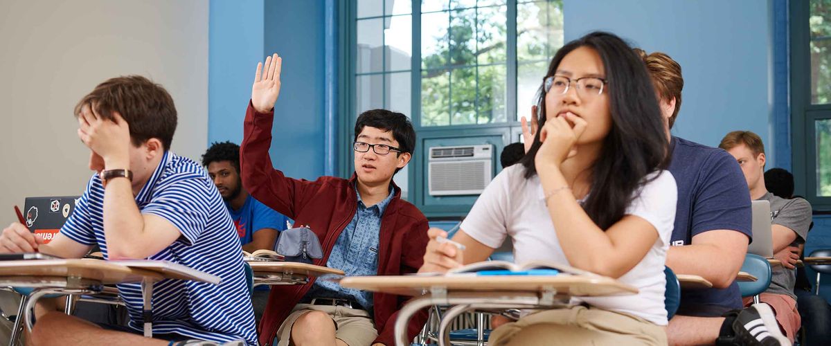 A BU Summer Term student seated at a desk and raising his hand in a classroom