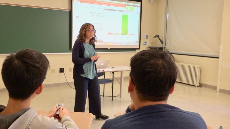 An instructor in a classroom with students and a screen of slides