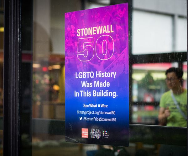 A blue and pink gradient flyer taped onto a window of BU building that reads: Stonewall 50 LGBTQ History was made in this building. See whaat it was: historyproject.org/stonewall50. The twitter logo followed by #BostonPrideStonewall50