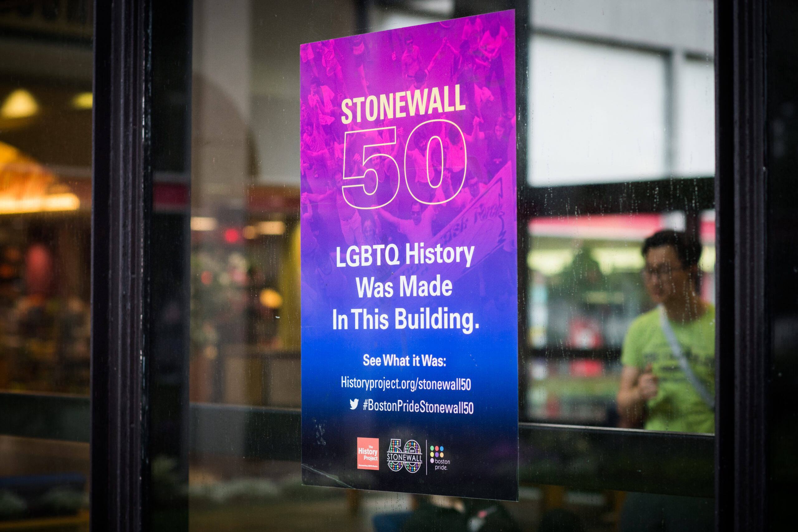 A blue and pink gradient flyer taped onto a window of BU building that reads: Stonewall 50 LGBTQ History was made in this building. See whaat it was: historyproject.org/stonewall50. The twitter logo followed by #BostonPrideStonewall50
