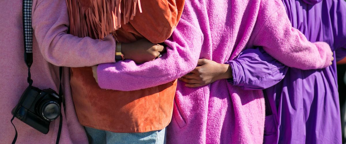 A zoomed in photo of a group of people with their backs facing the camera and their hands on each others backs. They are each wearing colorful clothing such was a pink jacket, an orange jacket, and a purple dress.