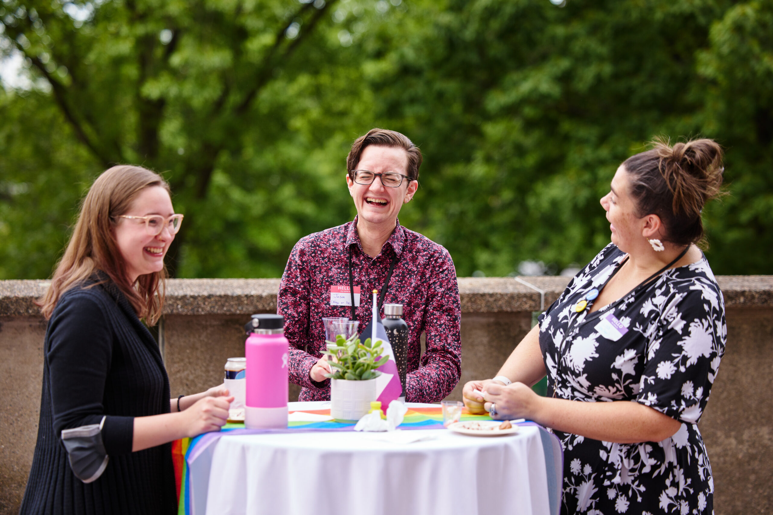 Three people standing round a cocktail table laughing. The person on the left has a pink water bottle on the table in front and there is a succulent plant as a center piece with rainbow table runners.