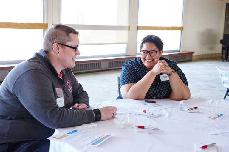 Two people at a round white table having conversation and smiling. There are pens and papers on the table in front of them.