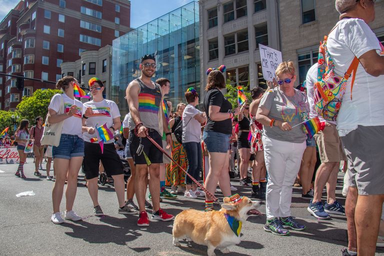 BU Pride parade with a variety of BU community members wearing rainbow garb on the road. In the center of the photo is a man wearing a rainbow tank top holding a corgi on a leash with a rainbow bandana.