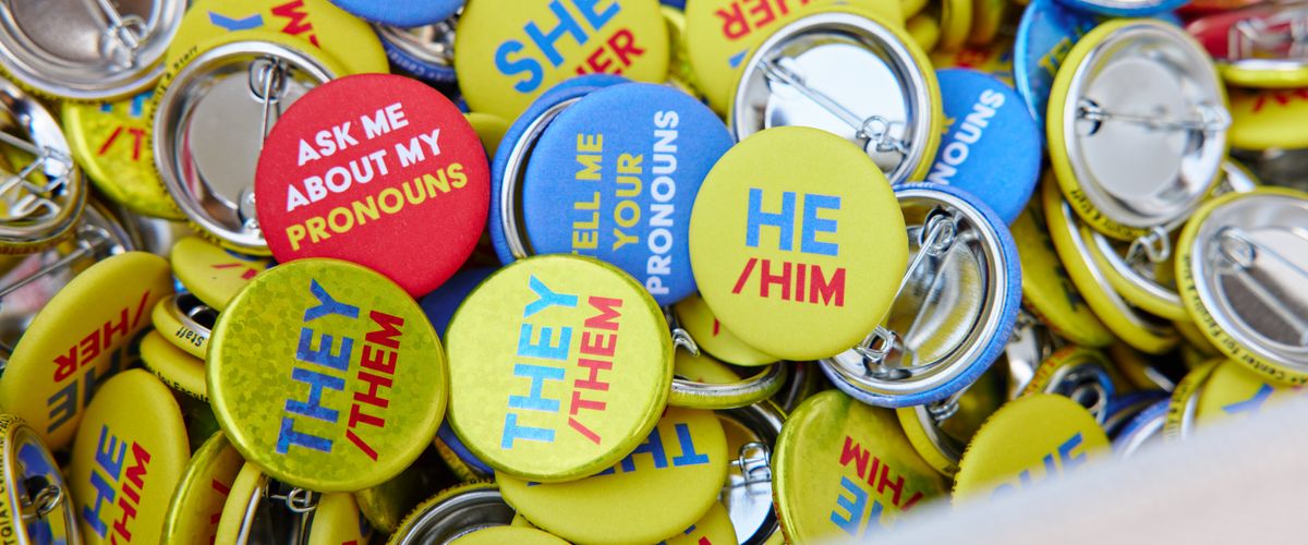 A close up photo of a variety of pronoun pins with the following pronouns: he/him, they/them, Ask me about my pronouns, She/Her, Tell me about your pronouns. The pins are jumbled up in a basket.