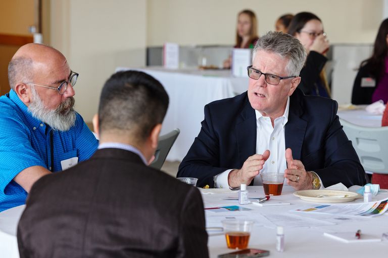 A person having a conversation at a round table with two others. The person is talking to the person directly across who has their back turned to the camera. Another person is beside the person speaking on the left side listening intently.