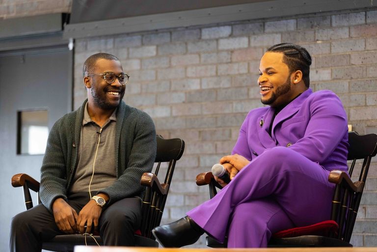 An image of WBUR reporter Quincy J. Walters and creative Jean Dolin. Quincy is a Black man with short buzzed black hair and short black stubble beard. He is wearing glasses, a green cardigan and a light gray polo shirt. He is smiling and looking over at Jean. Jean is a Black man with black braided hair. He is wearing a bright purple suit. He is smiling and looking out from where he is sitting.
