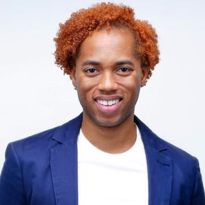A headshot of Ben Locke. Ben has dark skin, with short and curly ginger hair and is wearing a blue blazer and a white t-shirt.