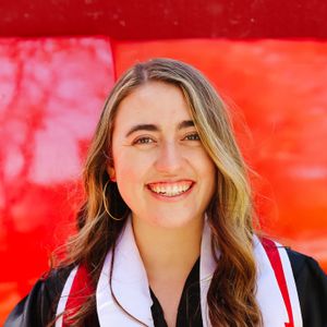 A headshot of Shelby Cattani. She has light skin, with long wavy blonde hair and is wearing a black gown wtih white and red stole. Shelby is standing in front of a red background.