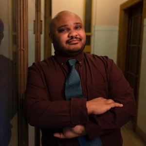 An image of Jam Hammond. Jam has medium dark skin with short black beard. He is wearing a dark red shirt with a blue tie and is posing with his arms crossed in front of him.