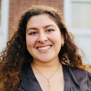 This is an image of Kayla. She has light skin, curly brown hair, and brown eyes. She is wearing a grey blouse and has gold earrings and a gold necklace.
