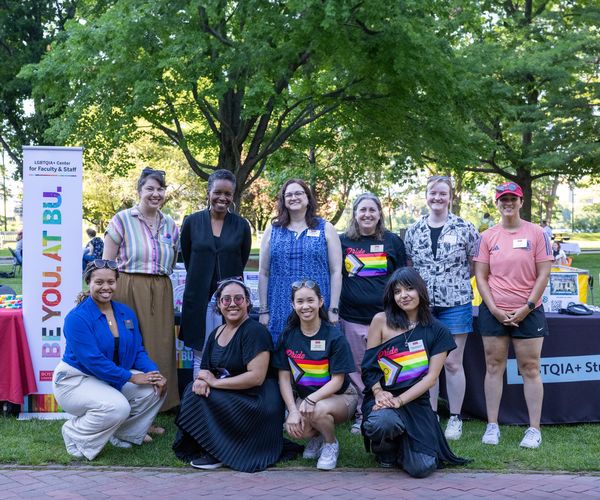 This is a group image of the Boston University LGBTQIA+ Centers teams celebrating at the Pride Fest 2024 event. There are five people standing and four people crouching at the bottom. They are all outdoors and posing in front of a big tree. To the left, there is a banner that reads "Be You. At BU."