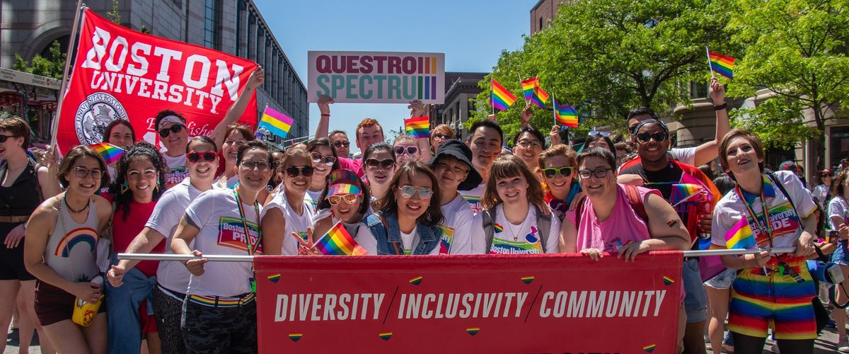 A group of people from the Boston University 2019 Pride Parade holding a red Boston University banner and small pride flags.