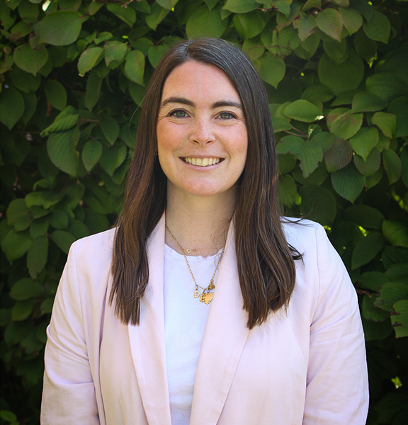 Headshot of Elizabeth Vassallo in a light pink blazer infront of a green bush