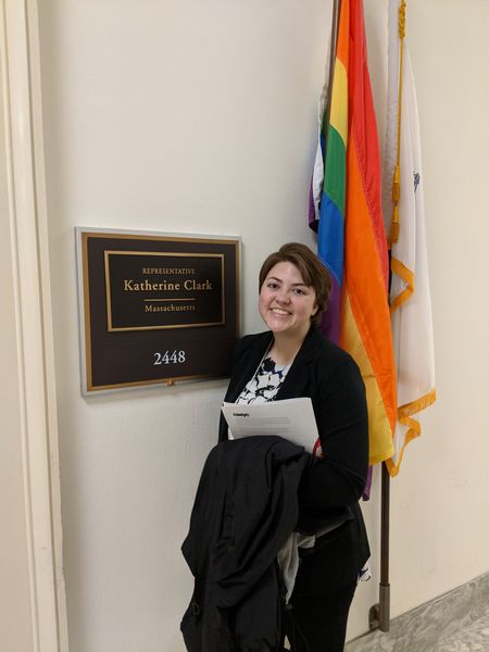 Krissy Hathaway (a short haired brunette in a suit) outside of Massachusetts Rep. Katherine Clark's office. There is a rainbow pride flag behind her.