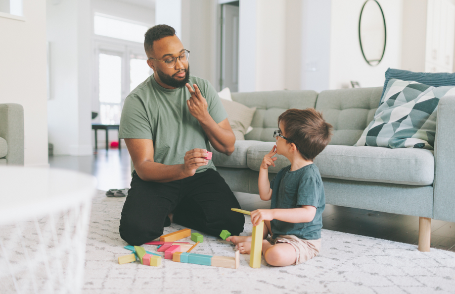 man sits on carpet in front of light blue couch using ASL to communicate to small child sitting next to him