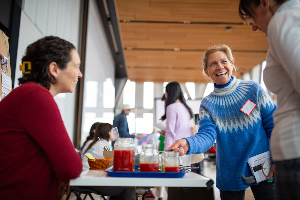 three women gathered around a booth with beakers filled with red liquid smiling