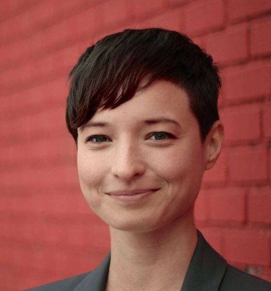 Shows Maya Bialik, head and shoulders against a red background, short brown hair and a green jacket