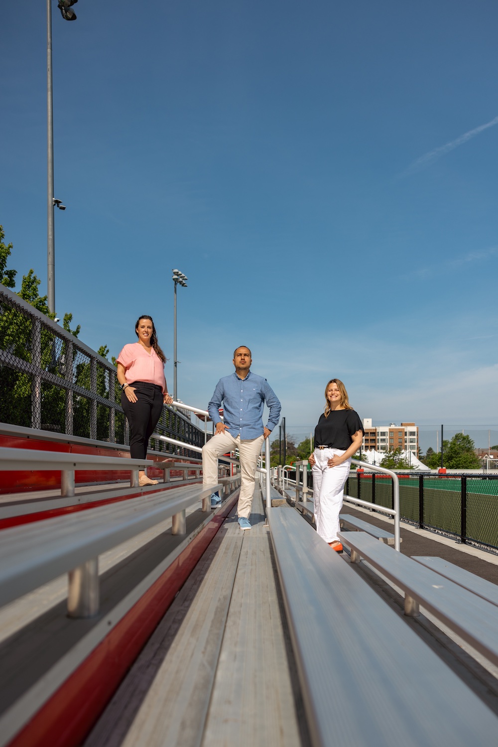 From L-R Anna Ward, Edson Filho, Carly Block standing and smiling on a set of bleachers