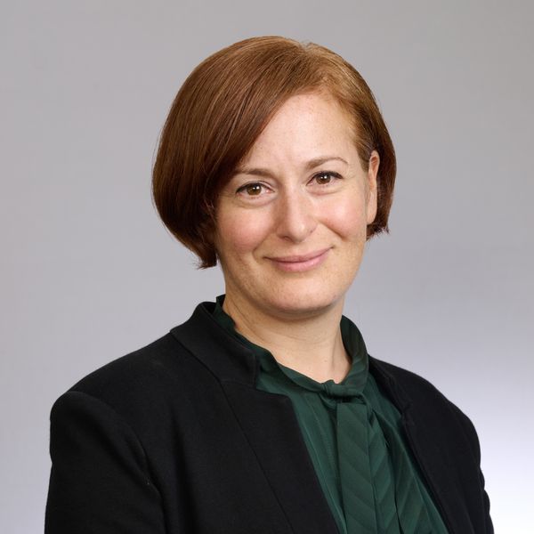Shows Ola Ozernov Palchik, who has short red hair and is wearing a green top and black jacket