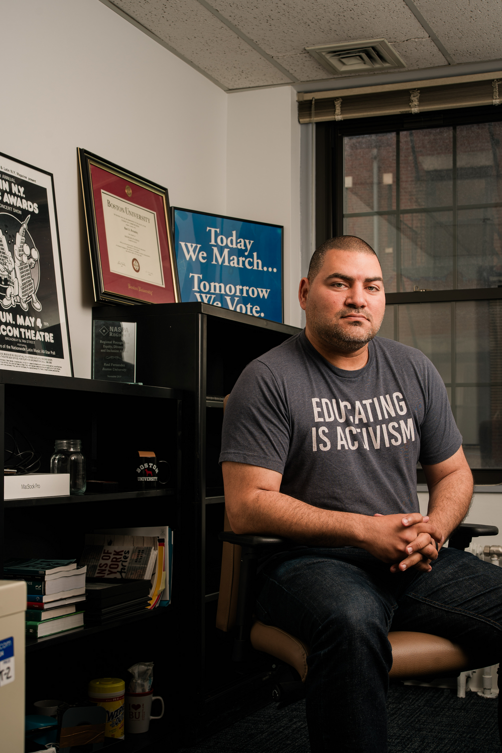 Raul Fernandez looking at the camera with a grey t-shirt reading "Educating is Activism" sitting in his office chair