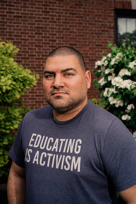Raul Fernandez looking at the camera with a grey t-shirt reading "Educating is Activism"