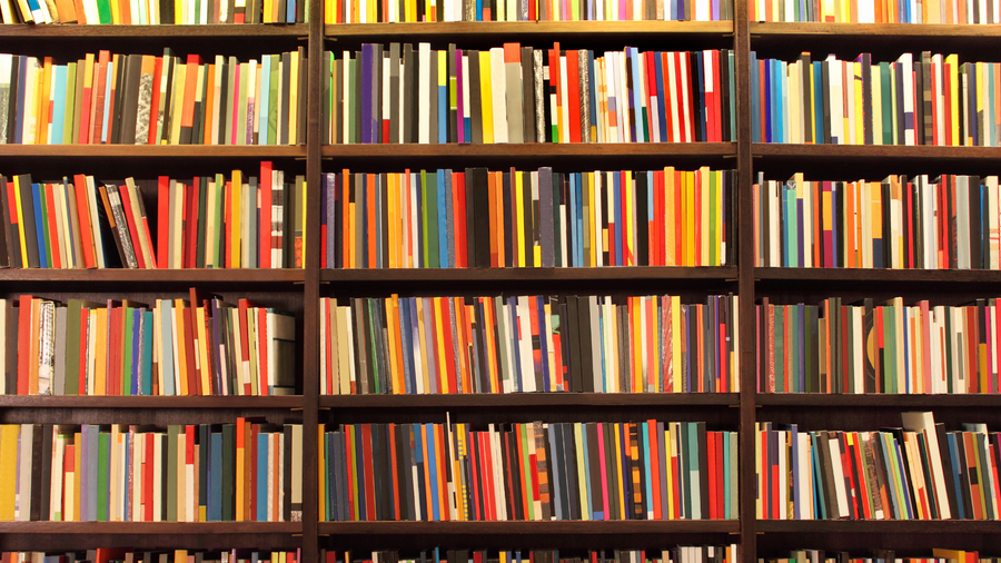A bookshelf filled with numerous colorful books arranged vertically. The books vary in size, thickness, and color, creating a vibrant and visually striking pattern.