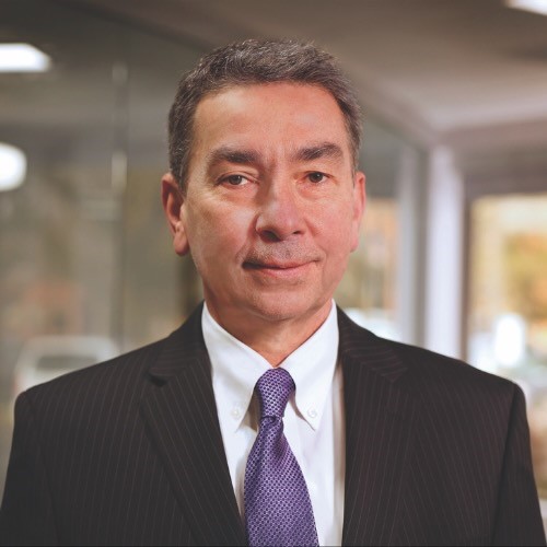 Shows a person with short hair wearing a dark jacket and purple tie