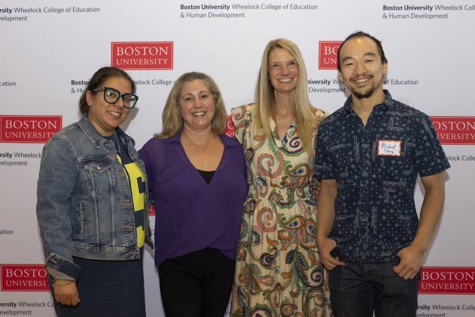 Shows four people standing in front of a step and repeat