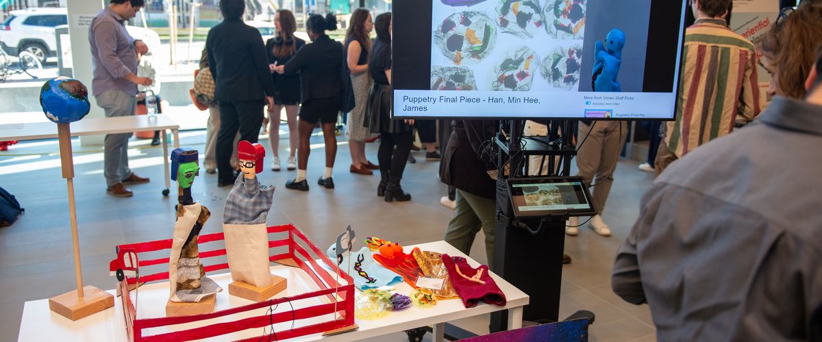 In a room, there's a table with puppet projects. A screen nearby displays puppet videos. Many students are present in the background.