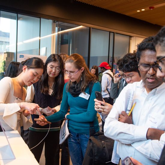 A photo from an event at Boston University displays interactions; a man observes as a teacher and a student examine something on a phone, while another person uses his phone to scan a QR code.