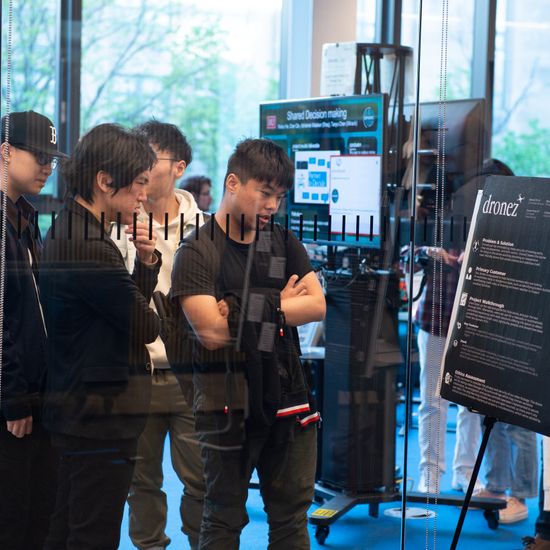 The picture features four students at a fair, each focused on different things. Some are studying a poster about drones, while another explores something of his own interest.
