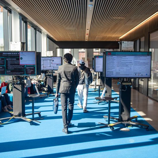 In a spacious room, screens are set up for an expo. Some students can be seen hanging around.
