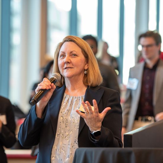 The picture shows a large room with a woman addressing the crowd with a microphone. It suggests an atmosphere of anticipation and learning.