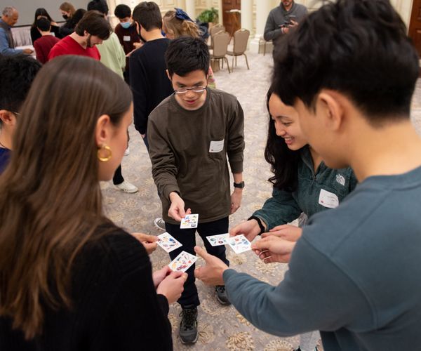 Five students stand in a small circle, engaged in a card game.